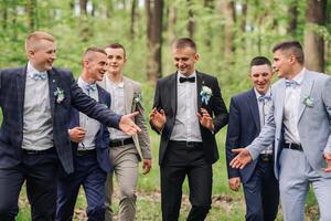 A group of men dressed in suits and ties are posing for a photo in a forest. Scene is lighthearted and fun, as the men are smiling and enjoying each other's company