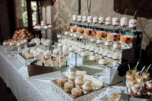 A table full of desserts and drinks with a white tablecloth. The desserts are arranged in a way that makes them look like they are on display. Scene is celebratory and inviting photo