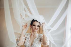 A bride is wearing a veil and standing in front of a curtain. She is smiling and looking at the camera photo