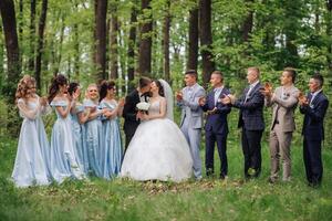 A group of people are standing in a field, with a bride and groom in the center. The bride is wearing a white dress and the groom is kissing her. The group is clapping and cheering for the newlyweds photo