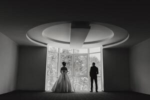 A man and a woman are standing in a room with a large window. The woman is wearing a white dress and the man is wearing a suit. Scene is one of anticipation and excitement photo