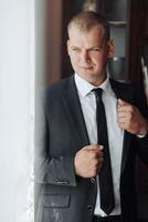 A man in a suit and tie is standing in front of a window, adjusting his tie. Concept of formality and professionalism, as the man is dressed in a suit and tie photo