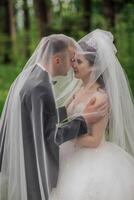 A bride and groom are standing in front of a white veil, with the bride's face partially hidden by the veil. The couple is dressed in formal attire, with the groom wearing a tie photo