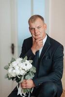 A man in a suit and tie is sitting on a chair with a bouquet of flowers in his lap photo