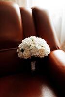 A bouquet of white flowers sits on a brown leather chair. The flowers are arranged in a vase and are placed on the arm of the chair. The chair is positioned in front of a window photo