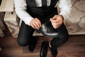 A man in a suit is putting on his shoes. He is sitting on a wooden floor. The shoes are black and shiny photo