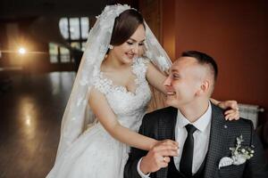 A bride and groom are posing for a picture in a room. The bride is wearing a white dress and a veil, while the groom is wearing a suit and tie. They are both smiling and seem happy to be together photo