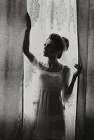 A woman in a wedding dress stands in front of a window. The image is black and white, giving it a classic and timeless feel. The woman's pose and the window's light create a sense of elegance photo