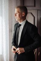 A man in a suit is standing in front of a window, adjusting his tie. Concept of formality and professionalism, as the man is dressed in a suit and tie. The window provides a view of the outside world photo