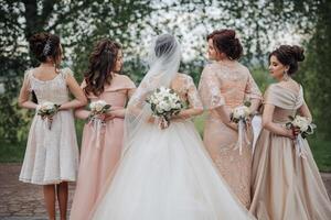 A group of women are dressed in wedding attire and holding flowers. They are standing in a field, with one woman wearing a white dress and another wearing a pink dress. The women are all smiling photo