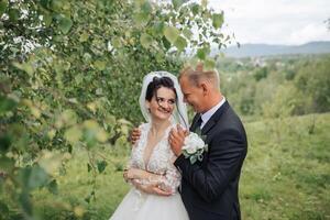 A bride and groom are standing in a field with trees in the background. The bride is wearing a white dress and the groom is wearing a suit. They are both smiling and seem happy photo