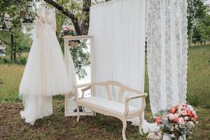 A white dress is hanging on a tree branch. A white bench is in front of a white curtain photo