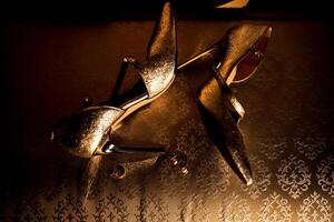 Two gold high heels are on a table. The heels are shiny and have a gold color. The table is made of wood and has a pattern on it photo