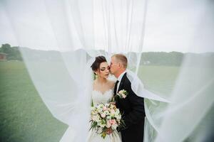 A bride and groom are kissing in front of a white curtain. The bride is wearing a white dress and the groom is wearing a black suit. The scene is intimate and romantic photo