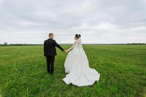 A bride and groom are walking in a field on a cloudy day. The bride is wearing a white dress and the groom is wearing a black suit. They are holding hands and seem to be enjoying their time together photo