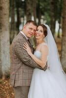 A bride and groom are posing for a picture in a forest. The bride is wearing a white dress and the groom is wearing a suit. They are both smiling and looking at the camera photo