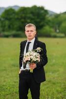 A man in a suit and tie is holding a bouquet of flowers. The image has a calm and serene mood, as the man stands in a field surrounded by nature. The flowers in the bouquet add a touch of color photo