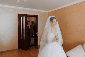 A bride is standing in front of a man, holding a bouquet of flowers. The man is wearing a suit and tie. The bride is smiling and looking at the camera. Scene is happy and celebratory photo