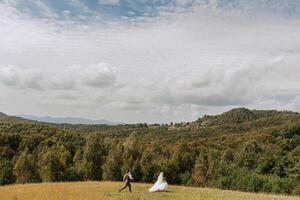 A bride and groom are running through a field with a beautiful view of mountains in the background. Scene is joyful and romantic photo