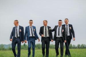 A group of five men are dressed in suits and ties, walking together in a field. Scene is one of camaraderie and friendship, as the men are enjoying each other's company and the outdoors photo