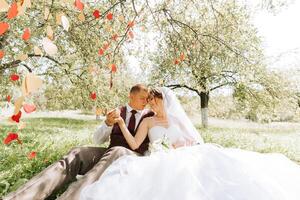 A bride and groom are sitting under a tree with red hearts hanging from the branches. They are holding hands and smiling at the camera photo