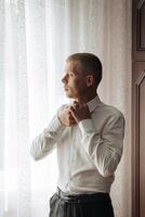 A man in a white shirt is getting ready to tie his tie. He is standing in front of a window, and the light coming in through the window is casting a warm glow on his face photo