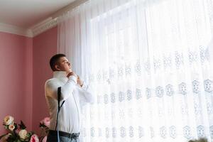 A man in a white shirt is getting dressed in front of a window. The curtains are white and have a pattern of squares. The man is wearing a tie and he is in a hurry photo