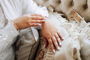 A woman is sitting on a couch with her hands on her lap. She is wearing a white robe and has her nails painted. The couch is covered in a floral pattern photo