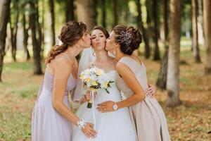 Three women are hugging and kissing each other in a park. One of them is wearing a wedding dress photo