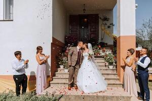 A bride and groom are kissing on a staircase in front of a group of people. The bride is wearing a white dress and the groom is wearing a suit. The people in the background are clapping photo