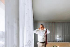 A man in a white shirt and gray pants is standing in front of a window, wearing a red tie. He is listening to music with his earbuds in photo