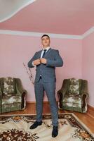A man in a suit is standing in front of two chairs. He is wearing a tie and adjusting it. The room has a pink wall and a green chair photo