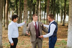 Three men are standing in a forest, one of them is wearing a suit and tie. They are all looking at something, possibly a camera or a piece of equipment. Scene is casual and relaxed photo
