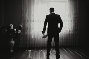 A man in a suit stands in front of a window with a bouquet of flowers in his hand photo