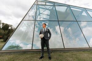 A man in a suit stands in front of a large glass building, holding a bouquet of flowers. Concept of elegance and sophistication, as the man is dressed in a formal attire photo