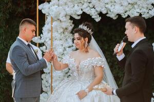 A bride and groom are standing under a white arch, with the bride wearing a tiara. The groom is clapping his hands in front of the bride photo