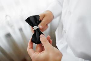A man is putting on a black bow tie with a diamond ring. Concept of elegance and sophistication, as the man is dressed in a formal outfit. The bow tie and ring add a touch of glamour to the scene photo