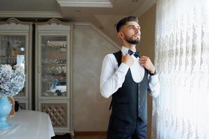 A man in a suit and tie is standing in front of a window, adjusting his tie. Concept of formality and professionalism, as the man is dressed in a suit and tie photo