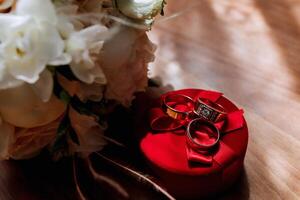A red box with three rings inside of it. The box is placed on a wooden table. The rings are gold and diamond-studded. The flowers in the background are white photo