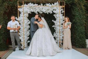 A bride and groom are kissing in front of a white arch. A man is standing behind them with a bouquet photo