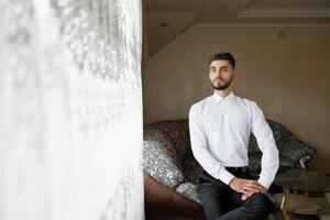 A man in a white shirt sits on a couch in a room with a window behind him. The room has a casual and comfortable atmosphere photo