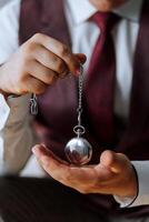 A man is holding a pocket watch in his hand. The watch is silver and has a chain around it. The man is wearing a red vest and a white shirt. Concept of nostalgia and elegance photo