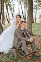 A bride and groom are posing for a picture in a forest. The bride is wearing a white dress and the groom is wearing a suit. They are sitting on a log in the woods photo