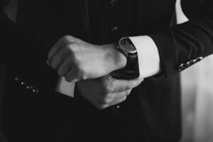A man is wearing a black suit and is looking at his watch. The watch is black and has a silver band. The man is in a hurry or is checking the time photo