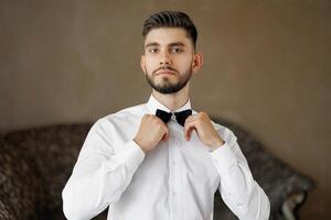 A man in a white shirt and black bow tie is getting ready to go out. He is standing in front of a brown wall photo