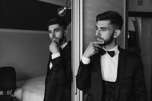 A man in a suit and tie is standing in front of a mirror, looking at his reflection. He is deep in thought, possibly contemplating his outfit or his day ahead. Concept of formality and professionalism photo