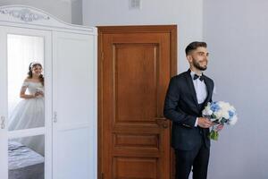 A man in a suit is standing in front of a door with a bride in a white dress peeking out from behind it photo