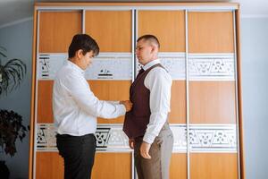 Two men are getting dressed up, one of them is wearing a vest. The man in the vest is adjusting his shirt while the other man looks on. Scene is casual and friendly photo