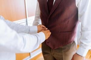 A man is getting dressed with a man helping him. The man is wearing a vest and a tie photo