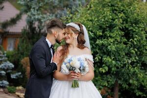 A bride and groom are standing in front of a house with a green bush behind them. The bride is holding a bouquet of flowers and the groom is kissing her on the cheek. Scene is romantic and happy photo
